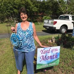 Woman in blue tie-dye top holding blue pinwheel beside “Yard of the Month – Unadilla, Georgia” sign and flower bed.