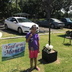 Child in purple shirt and pink shorts standing by “Yard of the Month – Unadilla, Georgia” sign near black lamppost and parked cars