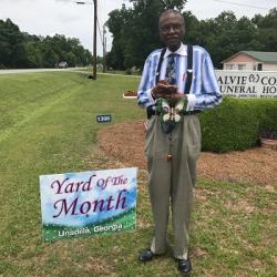 Person in striped shirt and suspenders standing next to “Yard of the Month – Unadilla, Georgia” sign on roadside lawn