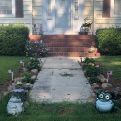 Yellow house entrance with white door, flower-lined concrete path, small frog statues, bushes, and American flags.