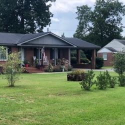 White brick house side with landscape plants, bird feeder on pole, chairs, and trees against green lawn.