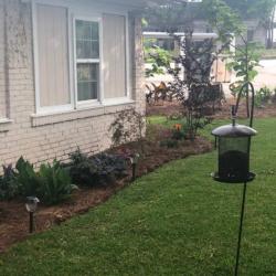 Brick ranch house with American flags on covered porch, surrounded by green lawn, flower beds, and trees under cloudy sky.